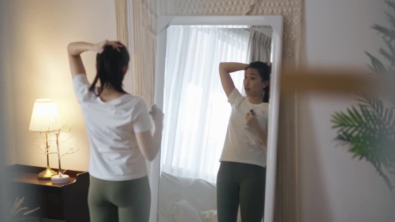 亚洲中国妇女系着她的头发准备瑜伽锻炼家庭锻炼在周末闲暇时间在镜子前视频下载