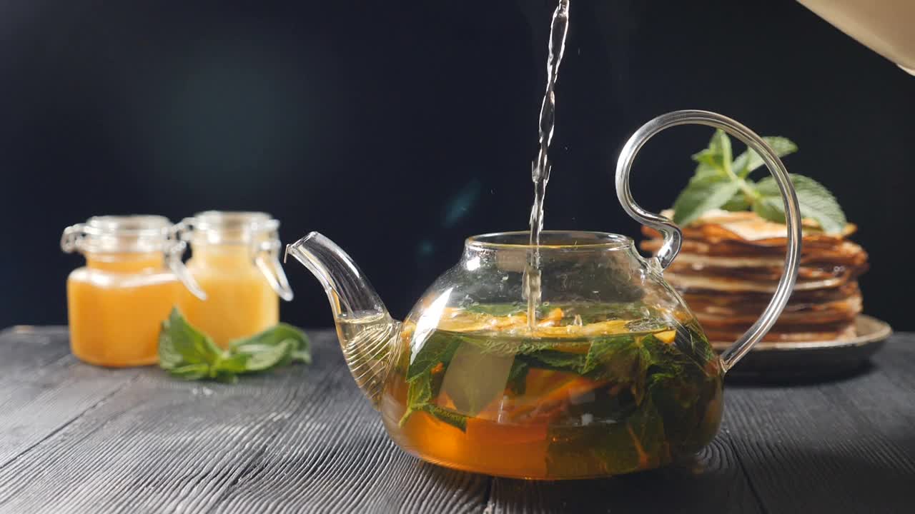 用沸水泡茶。背景是蜂蜜和一堆煎饼。在玻璃茶壶中向茶叶和橙片中加水。泡茶。抹茶茶道。全高清视频素材
