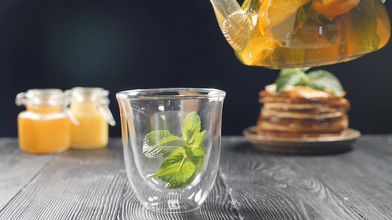 将橙汁、柠檬汁、青柠汁从透明的茶壶中倒入装有九香脂的玻璃杯中。以蜂蜜和薄饼为背景泡茶。慢镜头茶。全高清视频素材