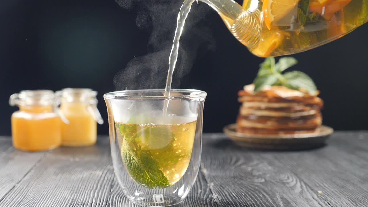 将橙汁、柠檬汁、青柠汁从透明的茶壶中倒入装有九香脂的玻璃杯中。以蜂蜜和薄饼为背景泡茶。慢镜头茶。全高清视频素材