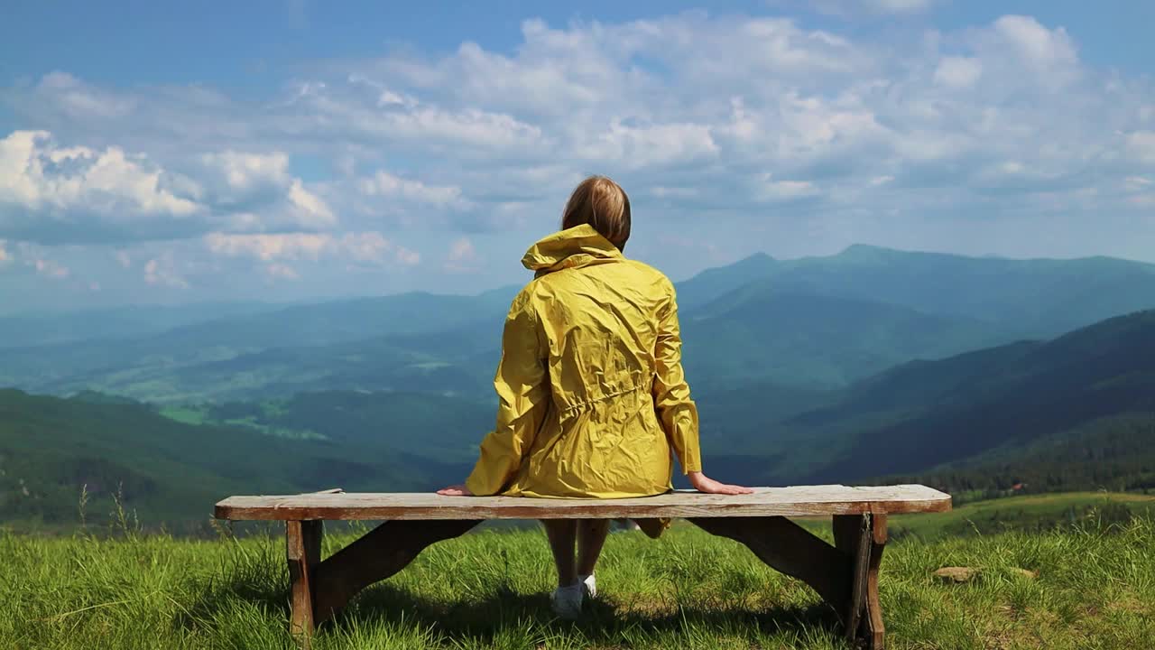 后视图年轻的女游客在明黄色夹克坐在路边的长椅上享受美丽的山景视频下载