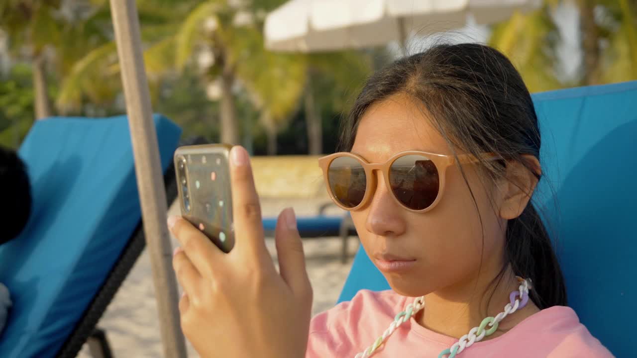 亚洲女孩使用智能手机，而放松在沙滩椅在早上假期与家人，生活方式的概念。视频素材