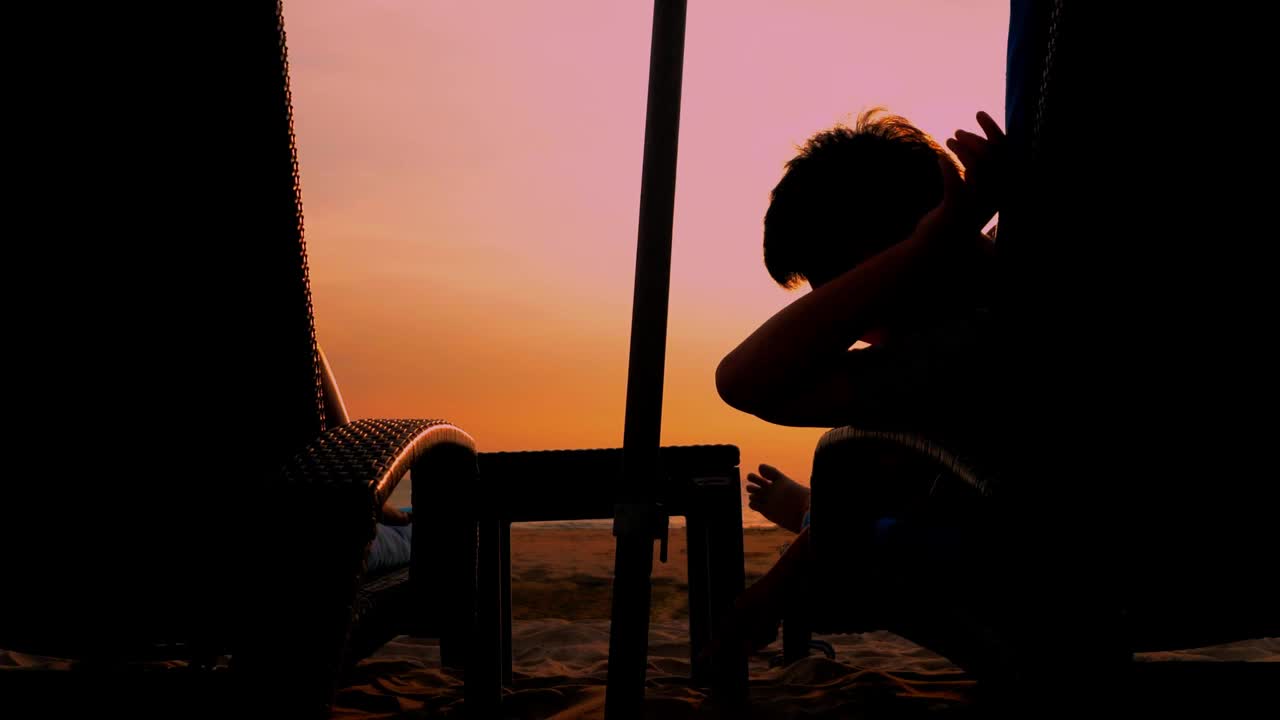 男孩坐在沙滩椅上画沙的剪影，以日出为背景，生活理念。视频下载