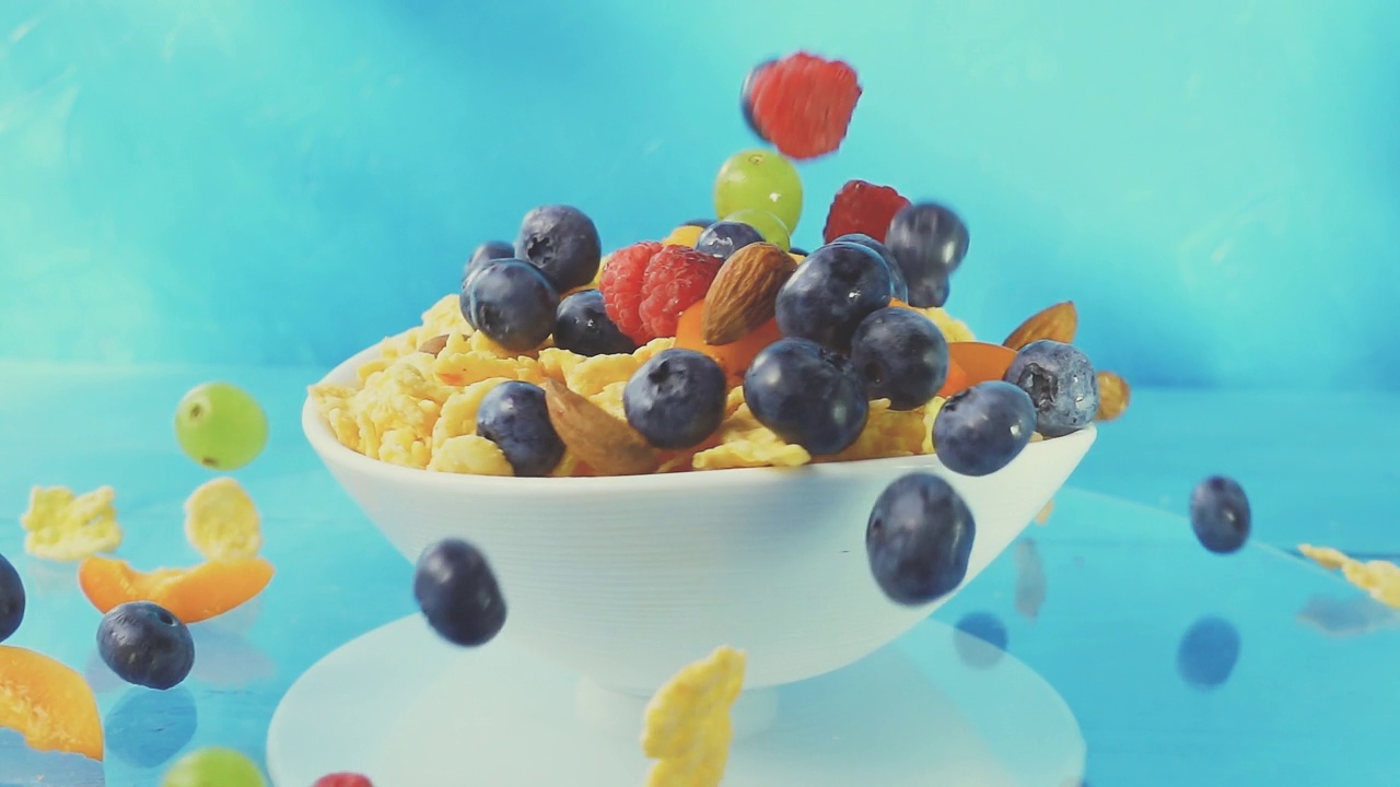 蓝莓、树莓、葡萄、杏仁和桃子的浆果落在一个旋转的白色盘子里，蓝色背景上放着黄色的玉米片。4k raw bright慢动作视频。视频下载