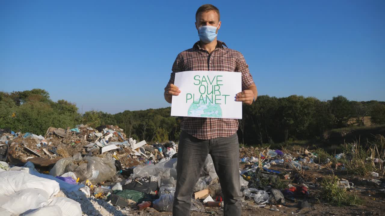 戴着医用防护口罩的年轻男子站在垃圾堆的背景下，对着镜头。男性环保人士手捧拯救地球的环保运动海报。视频下载