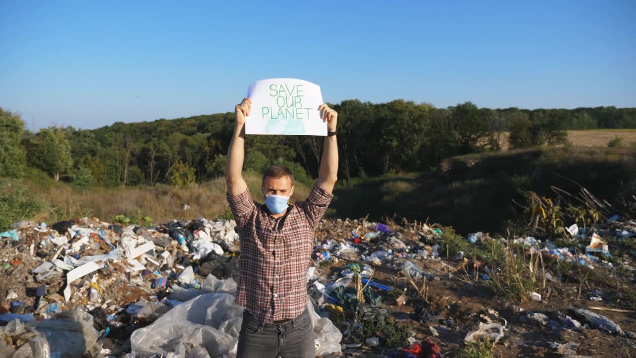 男性生态活动家头顶上高举拯救地球的海报，倡导自然保护。一名戴着防护面具的年轻人站在垃圾的背景下看着摄像机。视频下载