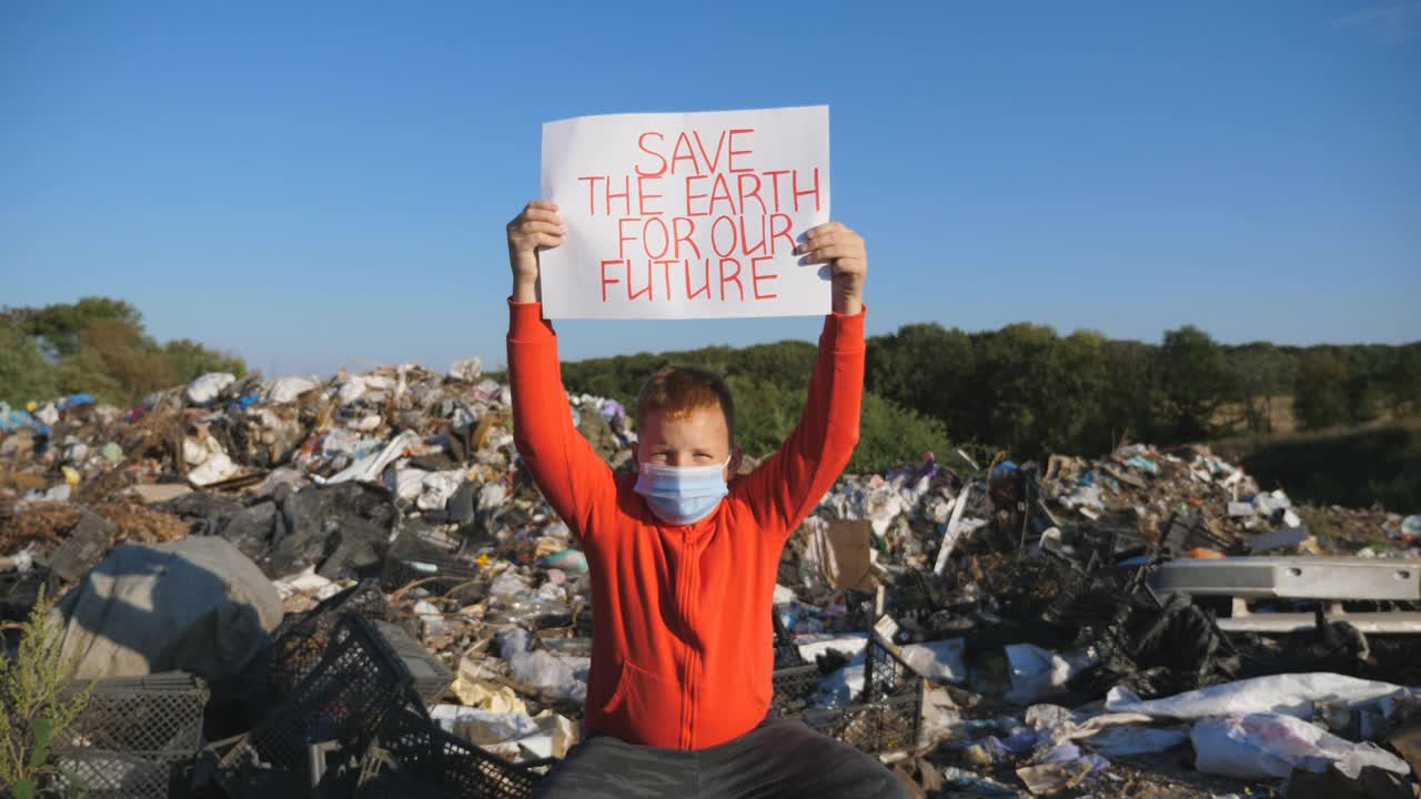 一个小男孩，头顶上举着一张拯救地球的海报，呼吁保护自然。戴着防护面具的小男性生态志愿者注视着相机，背景是垃圾填埋场。视频下载