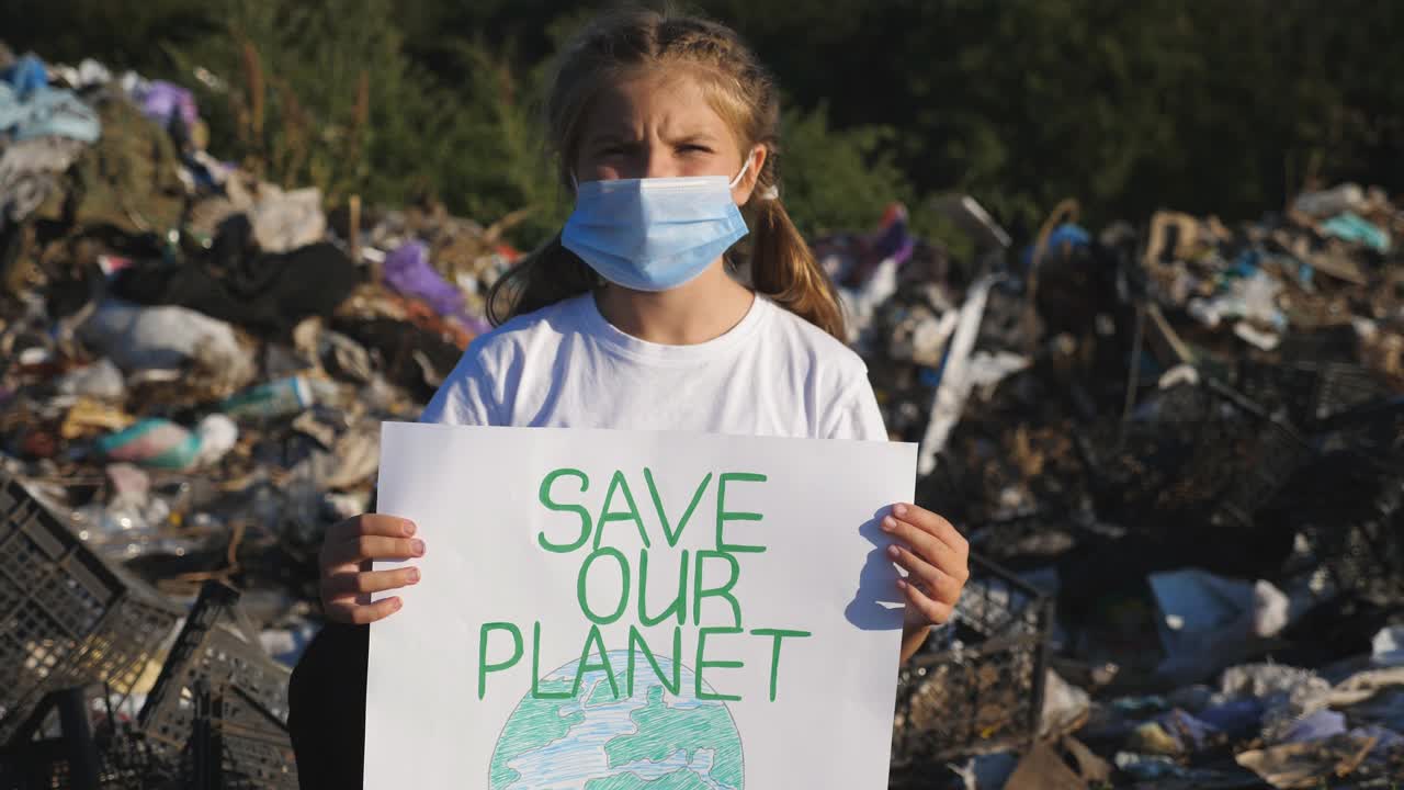小生态活动家的画像，手握环保运动标语，拯救地球。戴着防护口罩的小女孩在垃圾填埋场的背景下看着摄像机。近距离视频下载