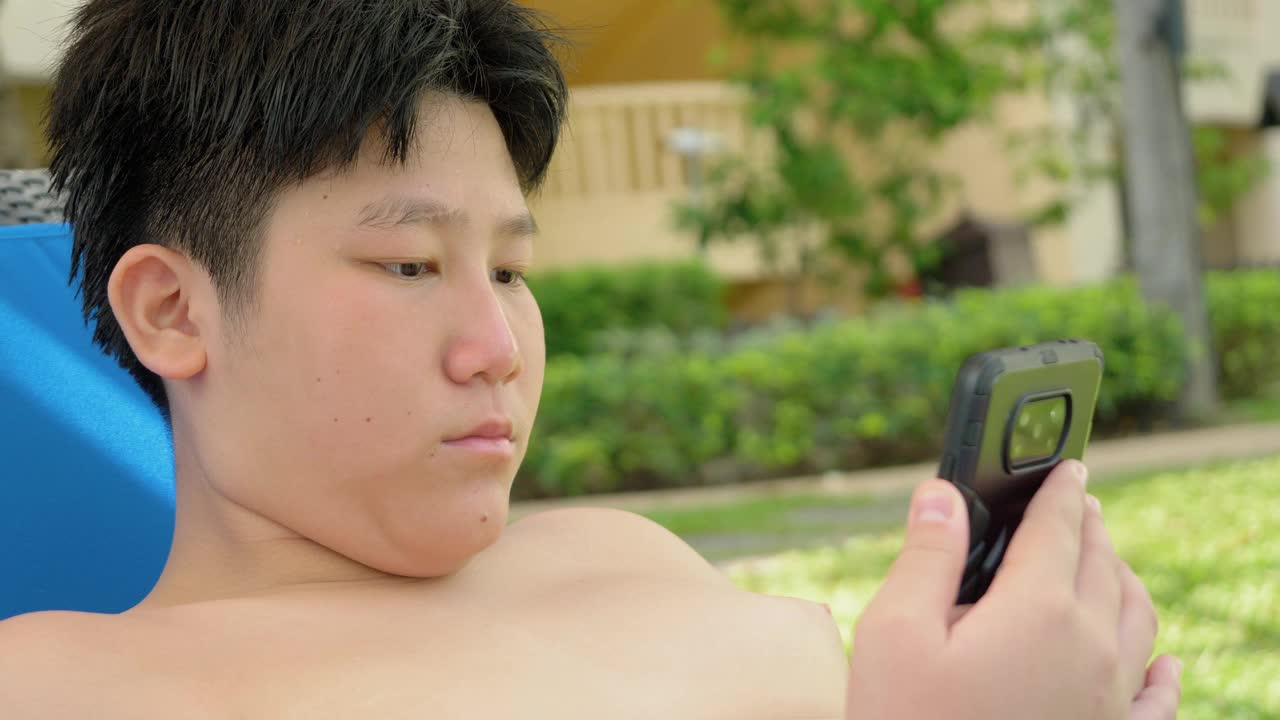 快乐的亚洲男孩使用智能手机，而放松附近的游泳池在酒店度假与家庭，生活理念。视频下载
