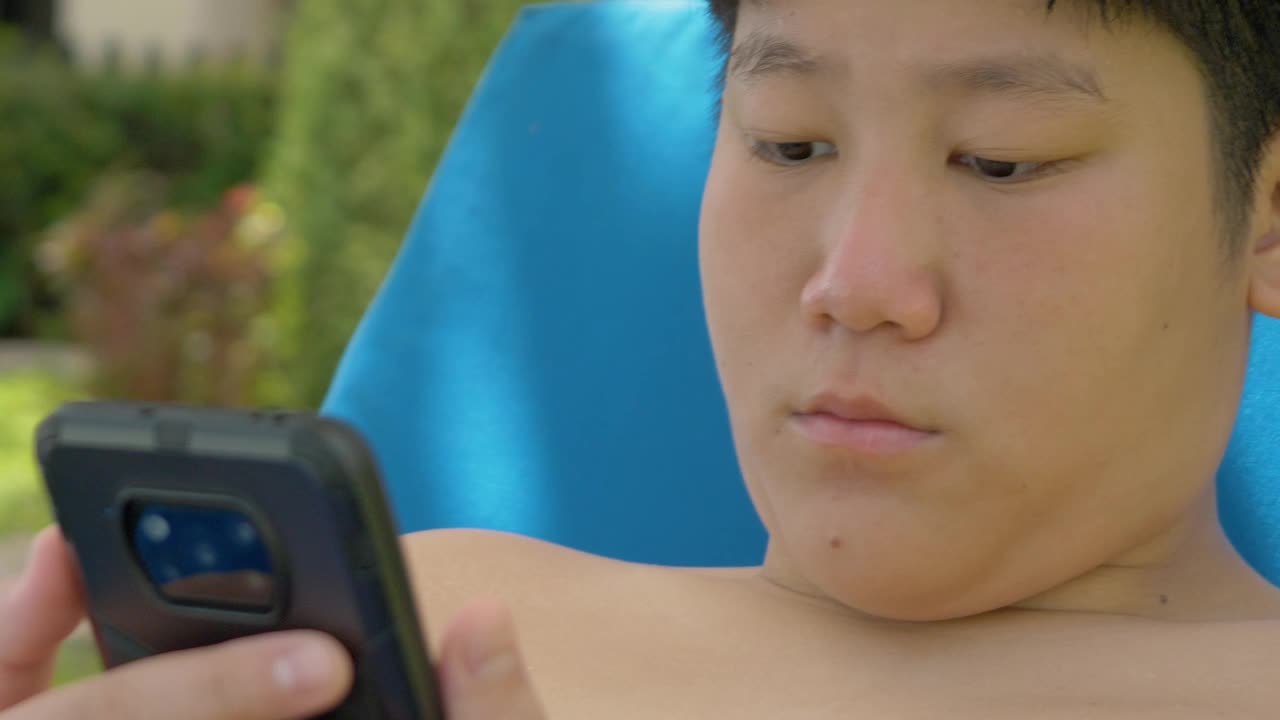 快乐的亚洲男孩使用智能手机，而放松附近的游泳池在酒店度假与家庭，生活理念。视频下载