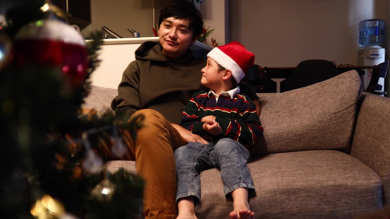 父亲和儿子坐在装饰着圣诞树的房间里的沙发上，一起玩耍。视频下载
