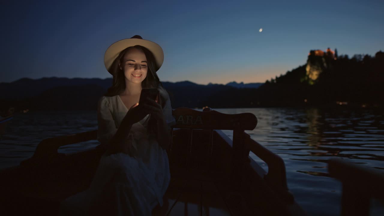 一名年轻女子在布莱德湖的一艘传统木船上使用智能手机视频下载