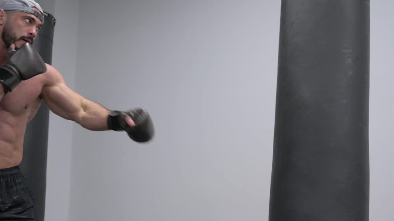 强壮的年轻大胡子踢拳白人男子在室内运动训练中戴着帽子和手套踢拳击袋用手和腿视频下载