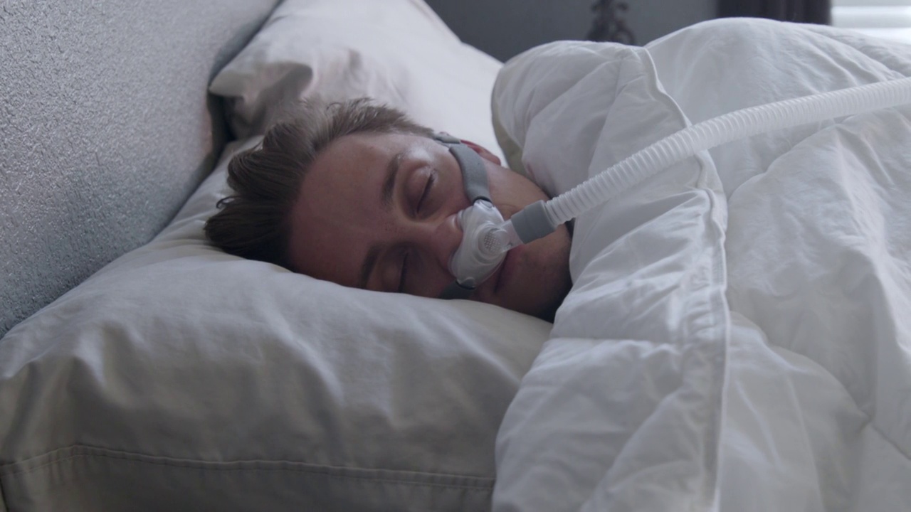 一个患有睡眠呼吸暂停的年轻人戴着CPAP口罩在床上侧睡视频下载