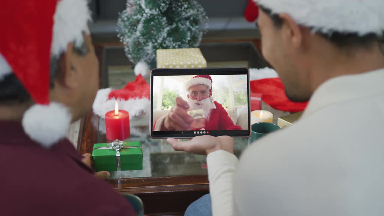 黑白混血的父亲和儿子戴着圣诞帽，用平板电脑进行圣诞视频通话，屏幕上有圣诞老人视频下载