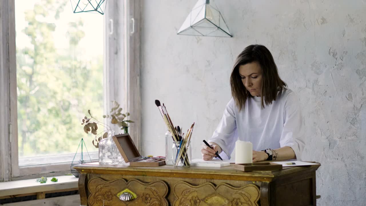 一个漂亮的黑皮肤女艺术家在她的木桌上用铅笔在纸上画画。4 k视频下载