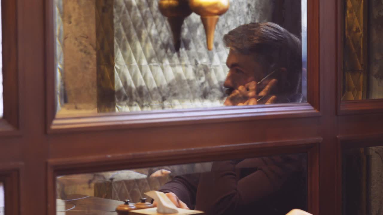 微笑的大胡子男人坐在桌旁，在镜子旁边讲电话视频素材