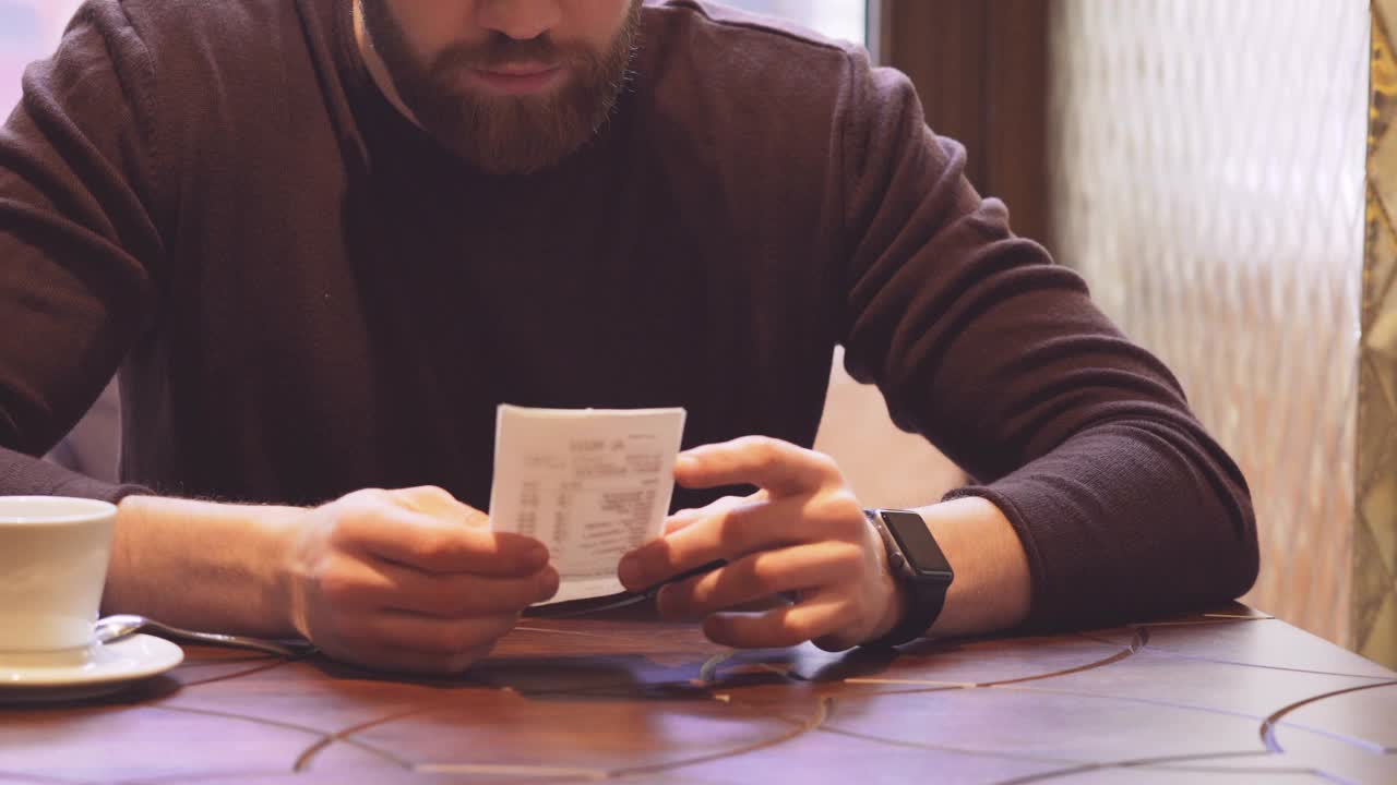 男子坐在桌子旁边，看着支票，用手表，通过无线支付技术支付了账单视频素材
