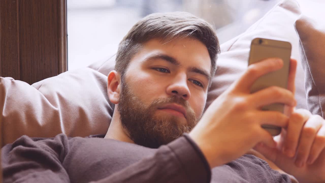 微笑大胡子男人躺在靠窗的沙发上用智能手机视频素材