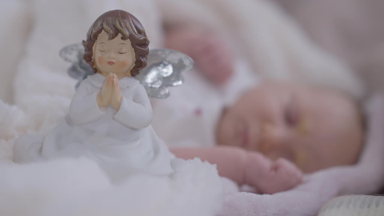 特写玩具祈祷天使和模糊无忧无虑的女婴睡在背景。放松的高加索婴儿休息在家里舒适的卧室室内。视频下载
