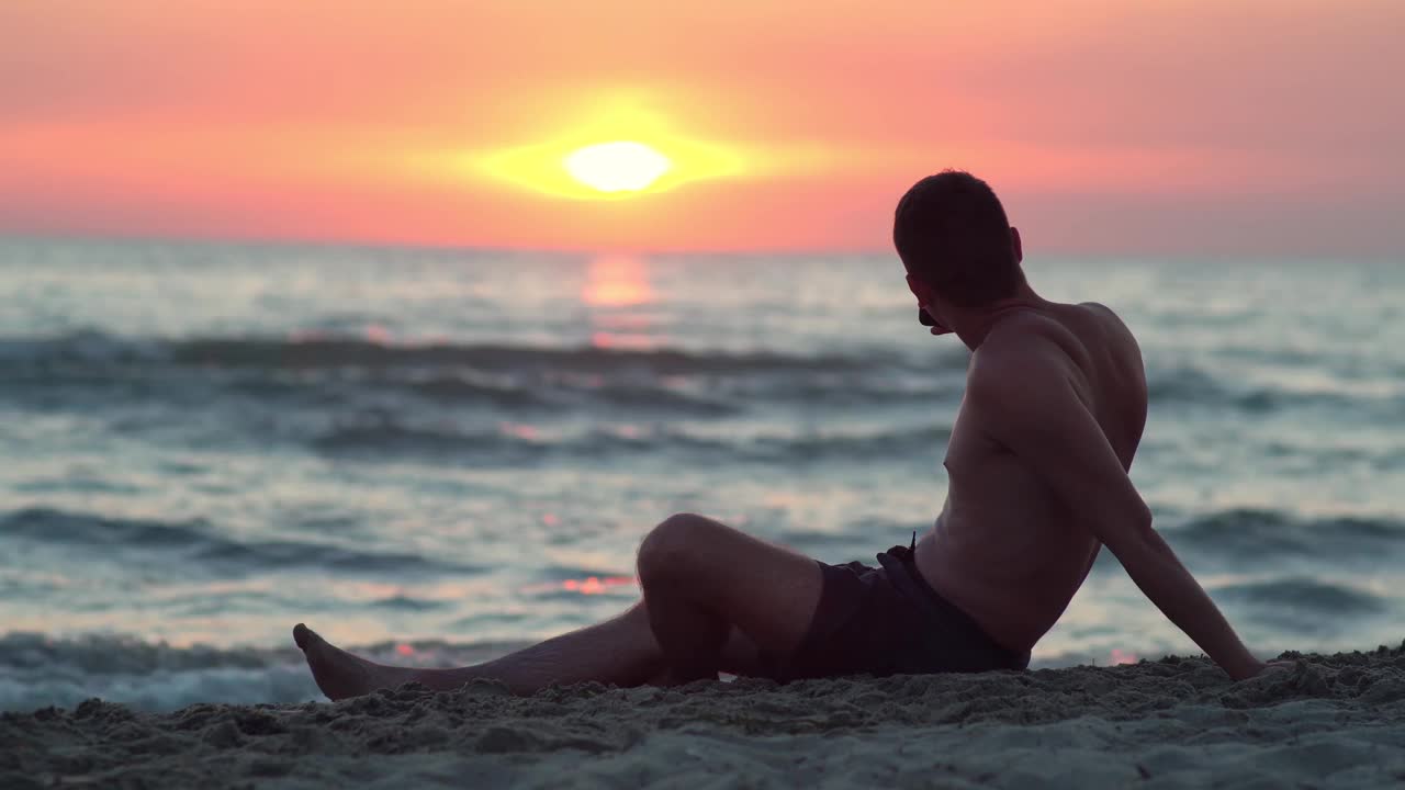 年轻人坐在热带沙滩上，用智能手机拍下美丽的海滩日落视频素材