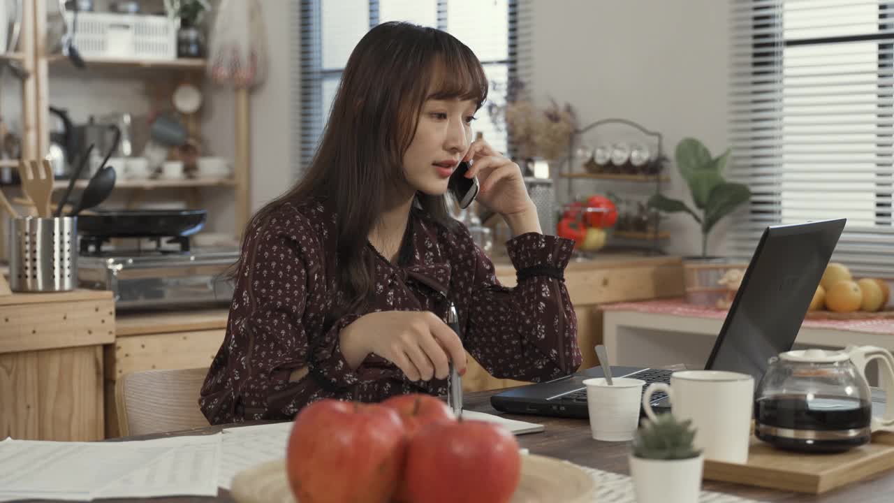 一位亚洲创意工作者在家里的餐厅里，抱着下巴，看着电脑沉思着，一边打字，一边打电话给她的客户视频素材