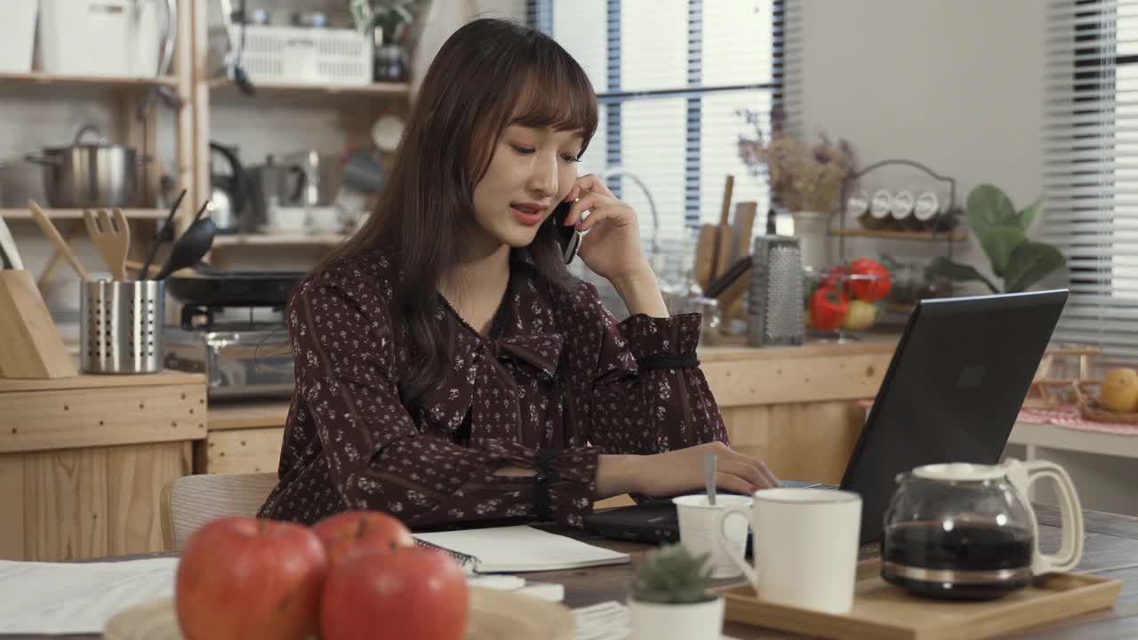 在家里工作的亚洲女性肖像指着笔记本电脑，与她的业务伙伴在餐桌上的电话讨论。视频素材