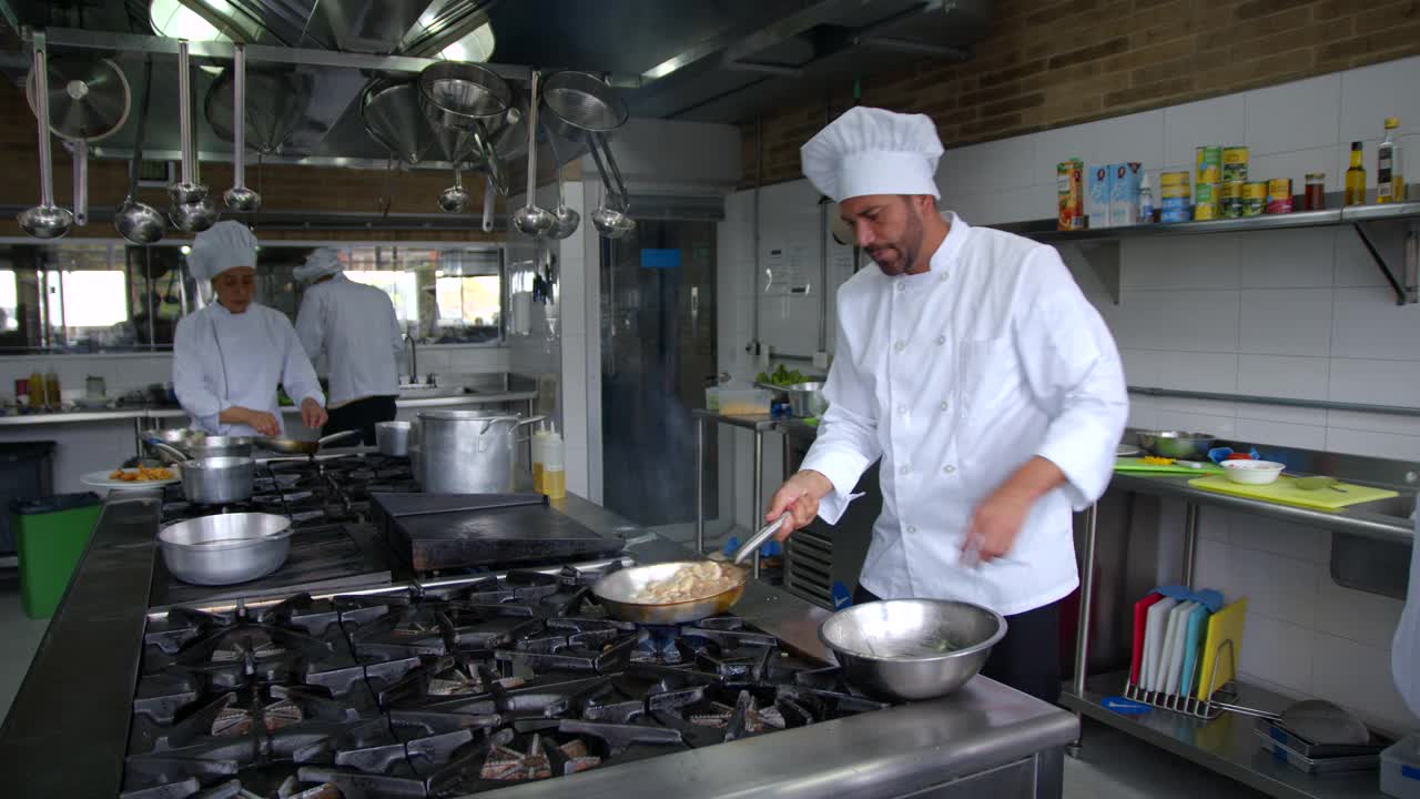 英俊的厨师sautéing在一个商业厨房的锅蔬菜，而其他两个副厨师的工作不同的饭菜视频下载