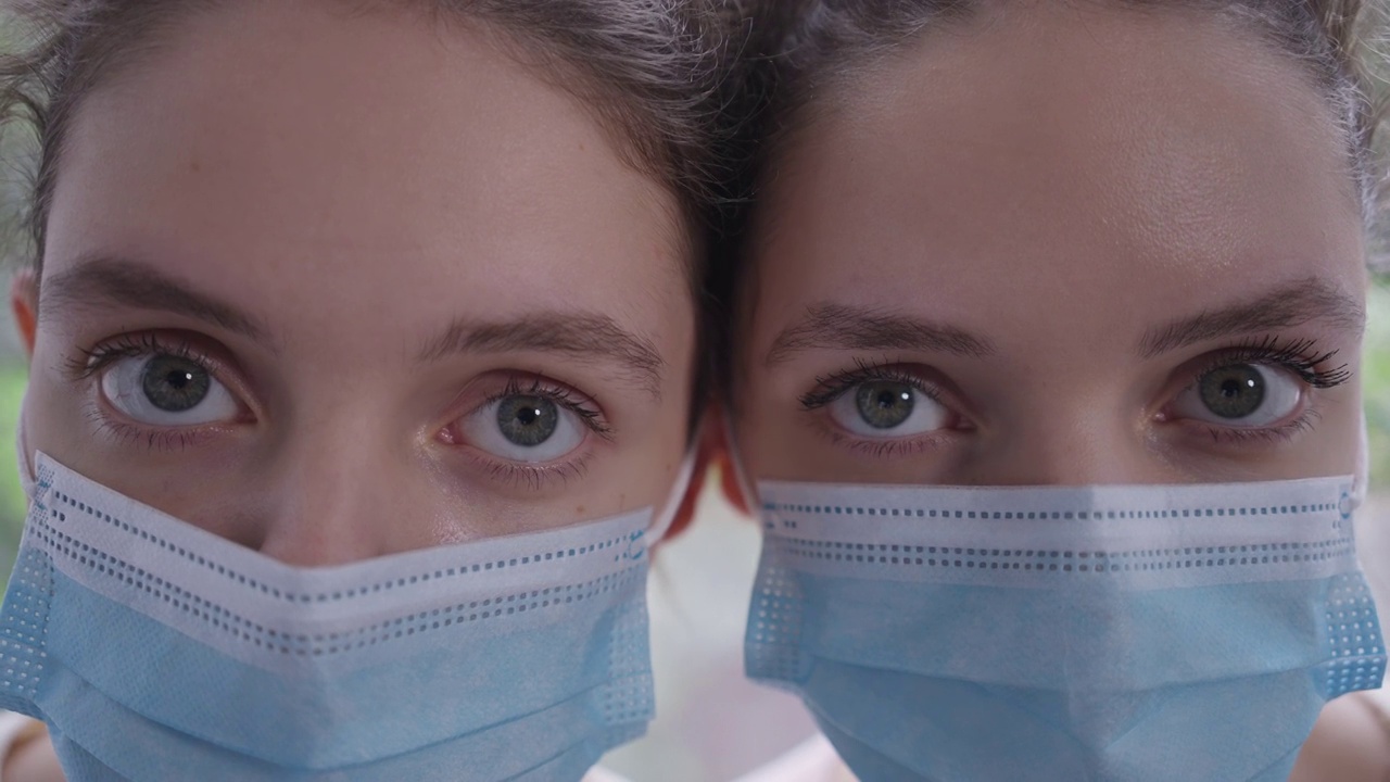 戴着冠状病毒口罩的同卵双胞胎姐妹的特写眼睛表情严肃地看着相机。苗条、年轻、美丽的白种女性因新冠疫情而被封锁的头像。视频下载