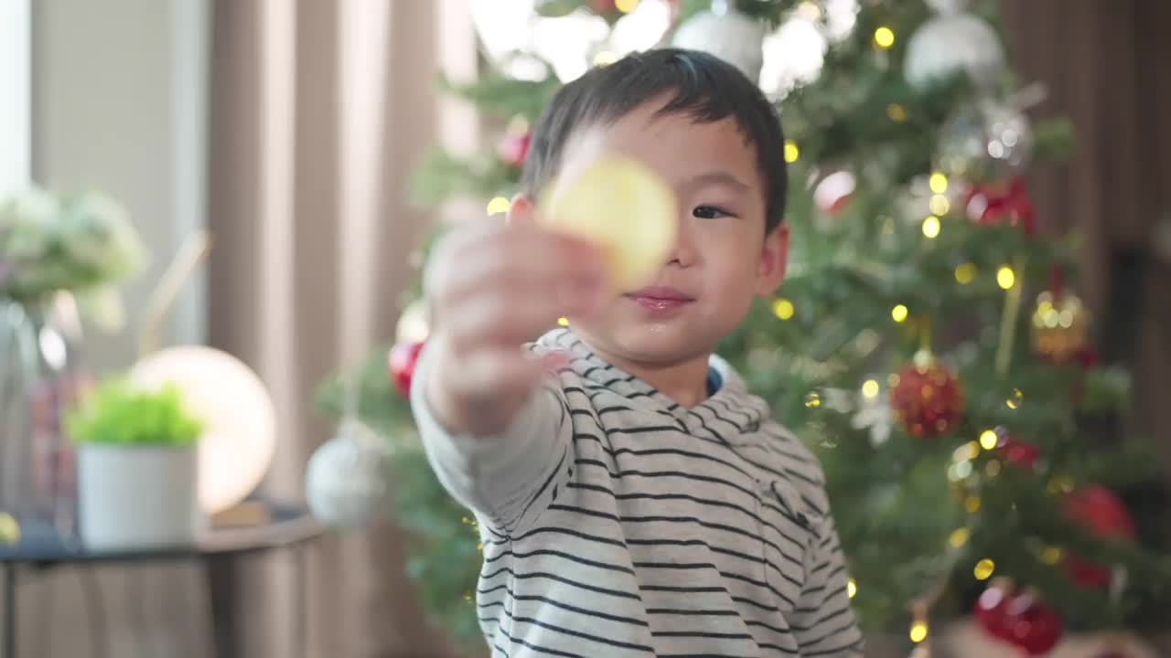 快乐而欢快的亚洲可爱清新的男孩笑着笑着快乐的时刻在圣诞树上装饰着闪亮的球和灯光，亚洲的孩子在客厅里玩着家里的圣诞树，圣诞节假期视频素材