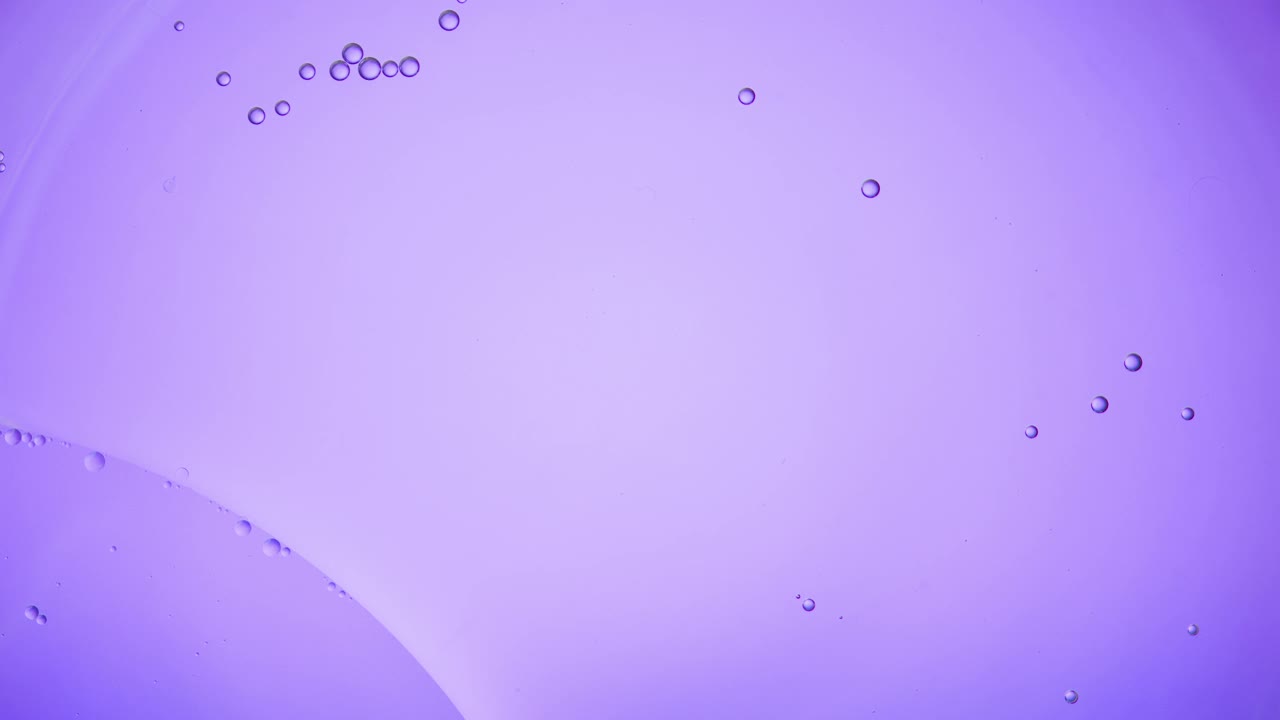 近距离和缓慢运动的头顶视图，倒清楚的油到一个池的水和填补与浮动油滴在一个背部照亮紫色的背景框架视频素材