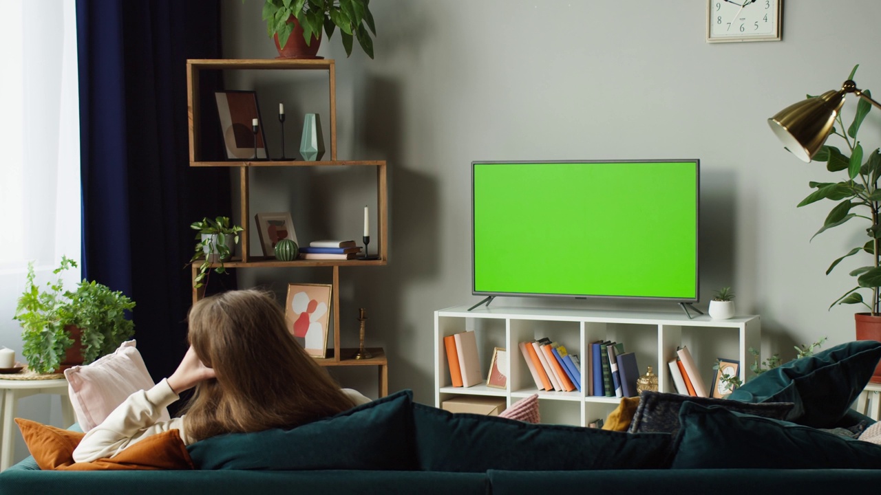 女人看电视与色度绿色屏幕。面目全非的女性坐在客厅沙发上，用遥控器切换电视频道。空余时间在家，放松身心视频素材