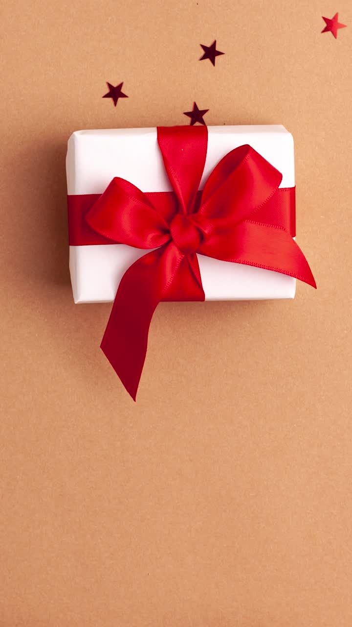 人的手在白色的毛衣拿走一个白色的纸礼物与红色缎子丝带蝴蝶结在棕色的背景与红色的星星形状。停止运动垂直动画圣诞假期概念平lay。视频素材