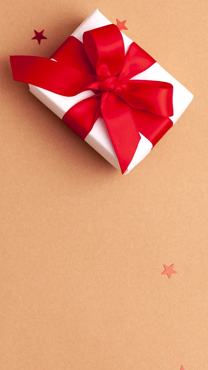 人的手在白色的毛衣拿走一个白色的纸礼物与红色缎子丝带蝴蝶结在棕色的背景与红色的星星形状。停止运动垂直动画圣诞假期概念平lay视频素材