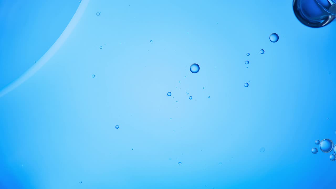 近距离和缓慢运动的头顶视图，倒清楚的油到一个池的水和填补与浮动油滴的框架在一个背景灯光蓝色背景视频素材