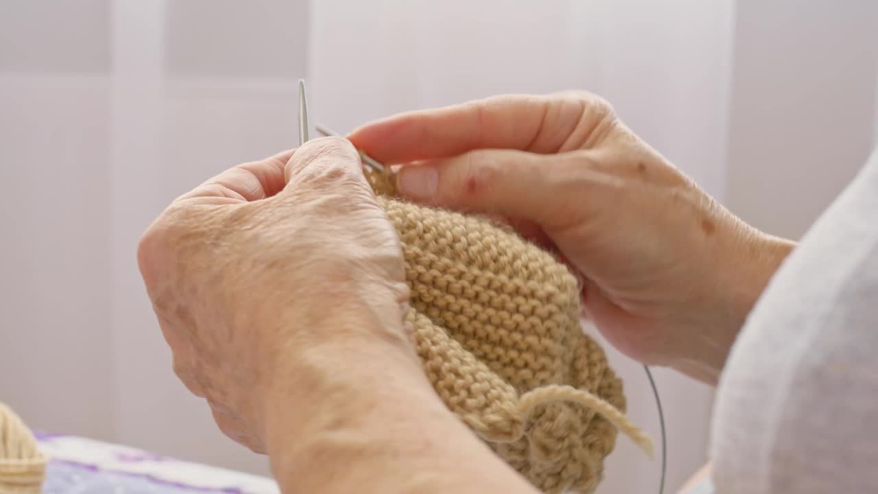老妇人拿着一根针织针和一件米色的针织衣服。老祖母们的最爱，在家自己动手织毛衣给孩子们手工织毛衣视频下载