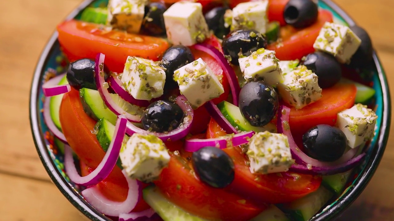 用碗盛着希腊沙拉，放在木板上。镜头移到一盘希腊沙拉上。夏日沙拉，有蔬菜、奶酪和橄榄。手动拍摄视频素材