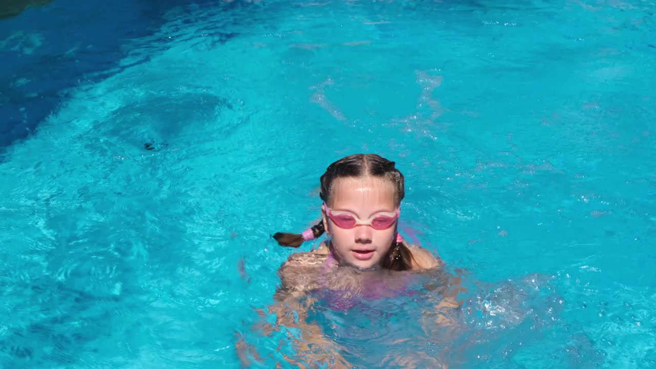 游泳池边戴着粉红色水杯的可爱女孩。女孩从水里冒出来视频下载