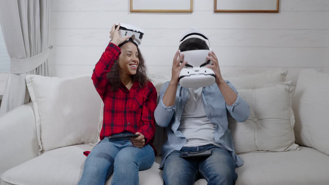 兴奋的西班牙年轻夫妇戴着虚拟现实头盔坐在沙发上，周末在家里用游戏机玩比赛游戏视频下载