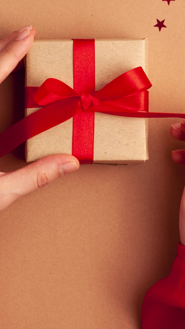 人的双手用红色的衣袖系好并解开棕色的纸礼物，用红色的缎带蝴蝶结在棕色的背景上有红色的星星形状。停止运动垂直动画圣诞节假期平lay视频素材