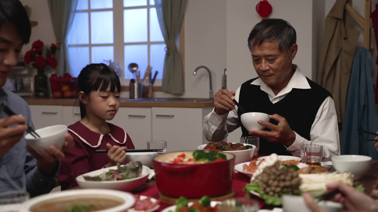 快乐的亚洲祖父与手势交谈，他礼貌的孙女把食物放进他的碗在中国新年前夕在家里的餐桌上视频购买