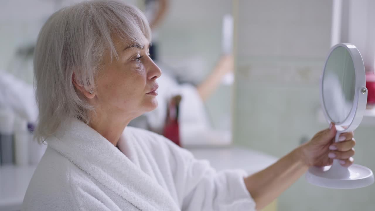 侧面看成熟的白人妇女看着手镜检查皱纹的脸和脖子。苗条的年长美丽退休人员肖像与老化在家里的浴室在室内视频素材