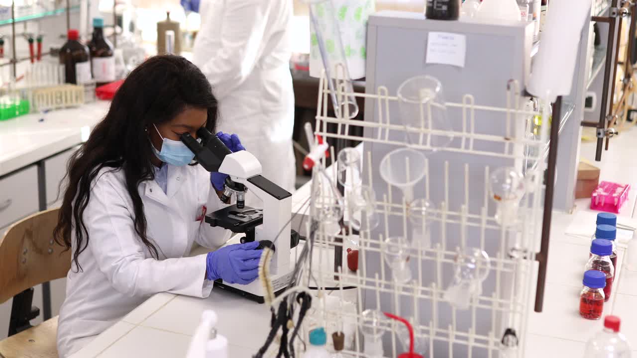 一位专注于微生物研究的女性微生物学家在她的同事工作的背景下检查微生物。视频下载