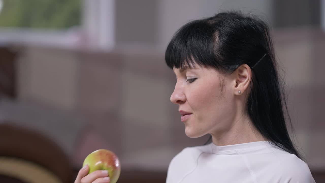 侧面看身材苗条的女人与黑头发咬嚼维生素苹果享受口味在慢动作。肖像美丽的白人自信的女士在家里吃水果。健康的生活方式。视频素材