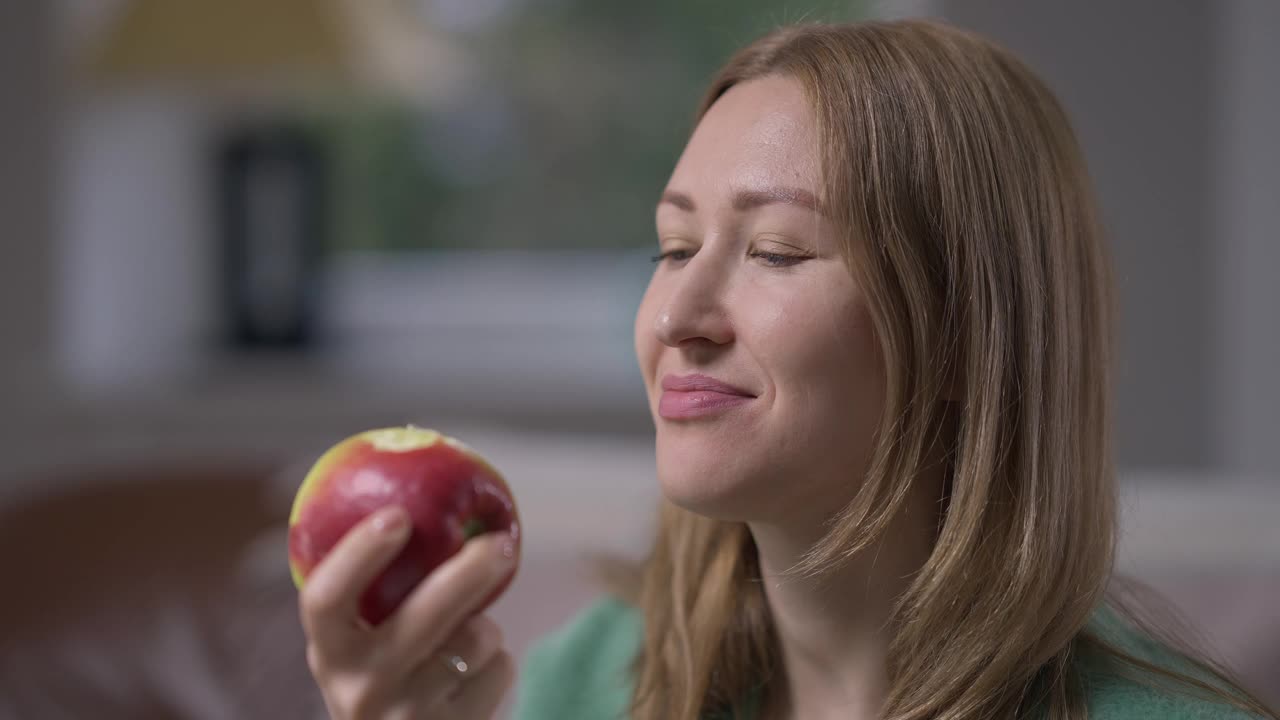 近距离微笑的女人吃健康的苹果看着相机微笑。积极自信的白种人苗条有吸引力的女士享受维生素水果的味道在室内摆慢动作。视频素材
