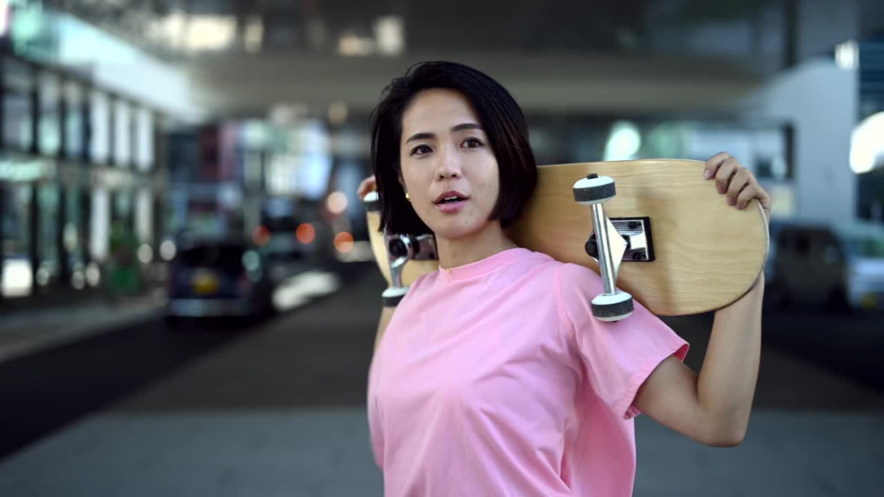 一名亚洲女子站在立交桥下，头后举着滑板。视频下载