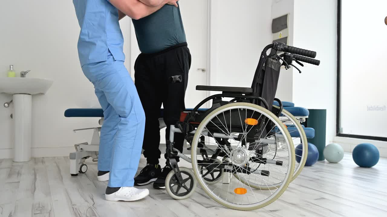 康复医院男物理治疗师，帮助坐轮椅的残疾人站起来。高质量4k镜头视频下载