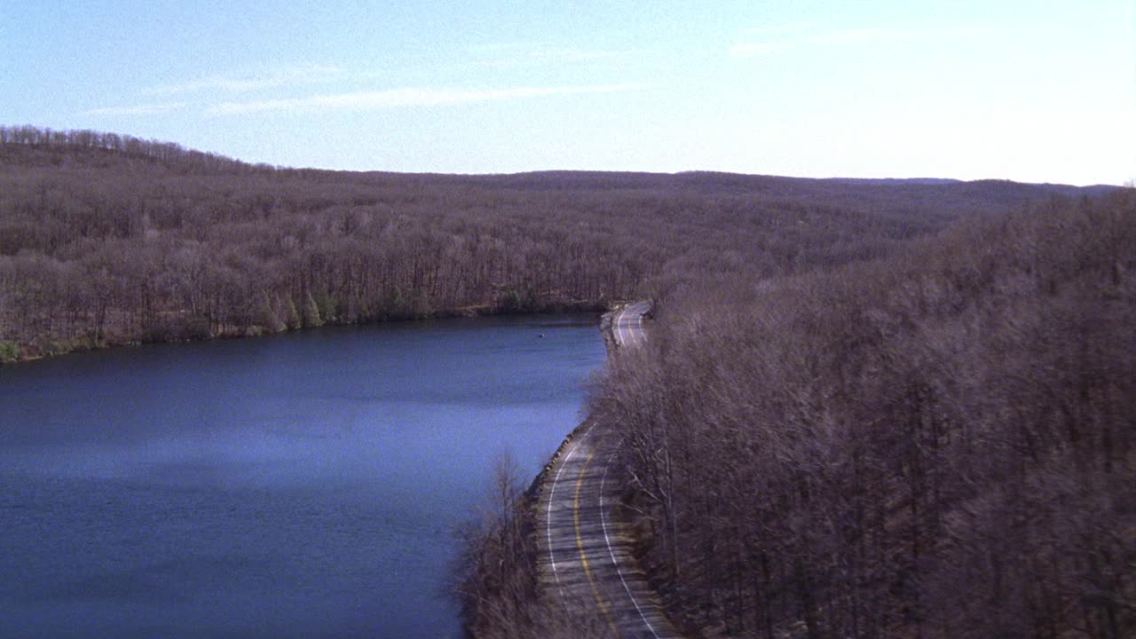 航空跟踪1998年福特金牛座汽车在两车道沿海公路。河对面山上光秃秃的树枝。可能是深秋或初冬。农村地区。视频下载