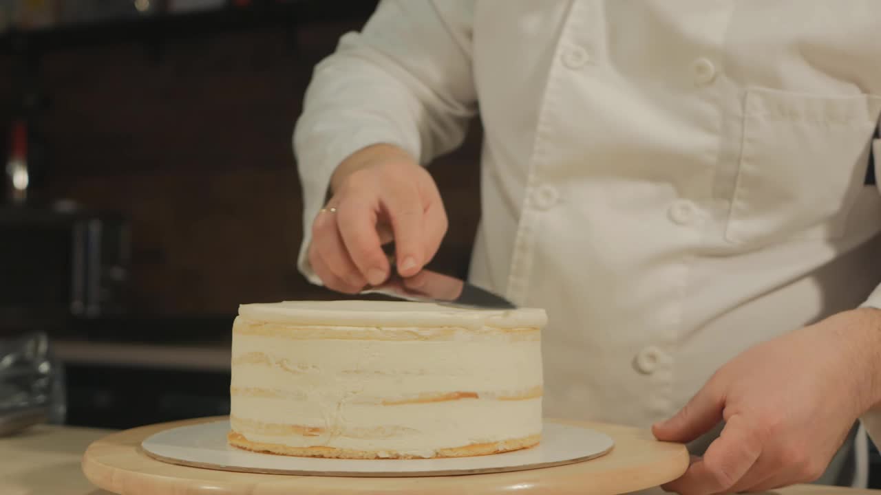 一名欧洲男子糕点厨师正在制作蛋糕准备表格。他用一把锋利的刀把蛋糕上的奶油磨平了。制作甜点视频下载