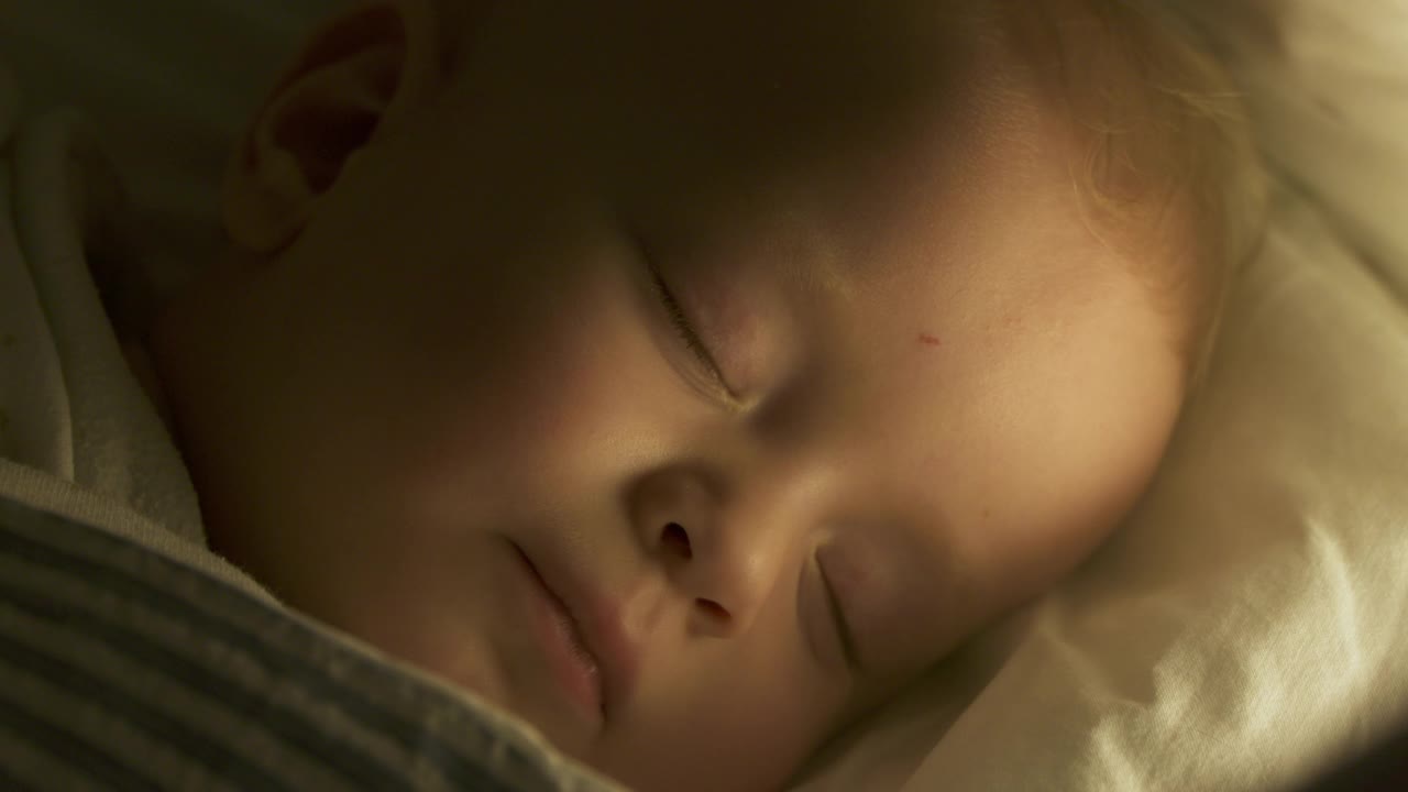 婴儿襁褓躺在婴儿床，9个月的男婴睡在夜灯下。9个月大的白人小孩的脸特写。视频下载